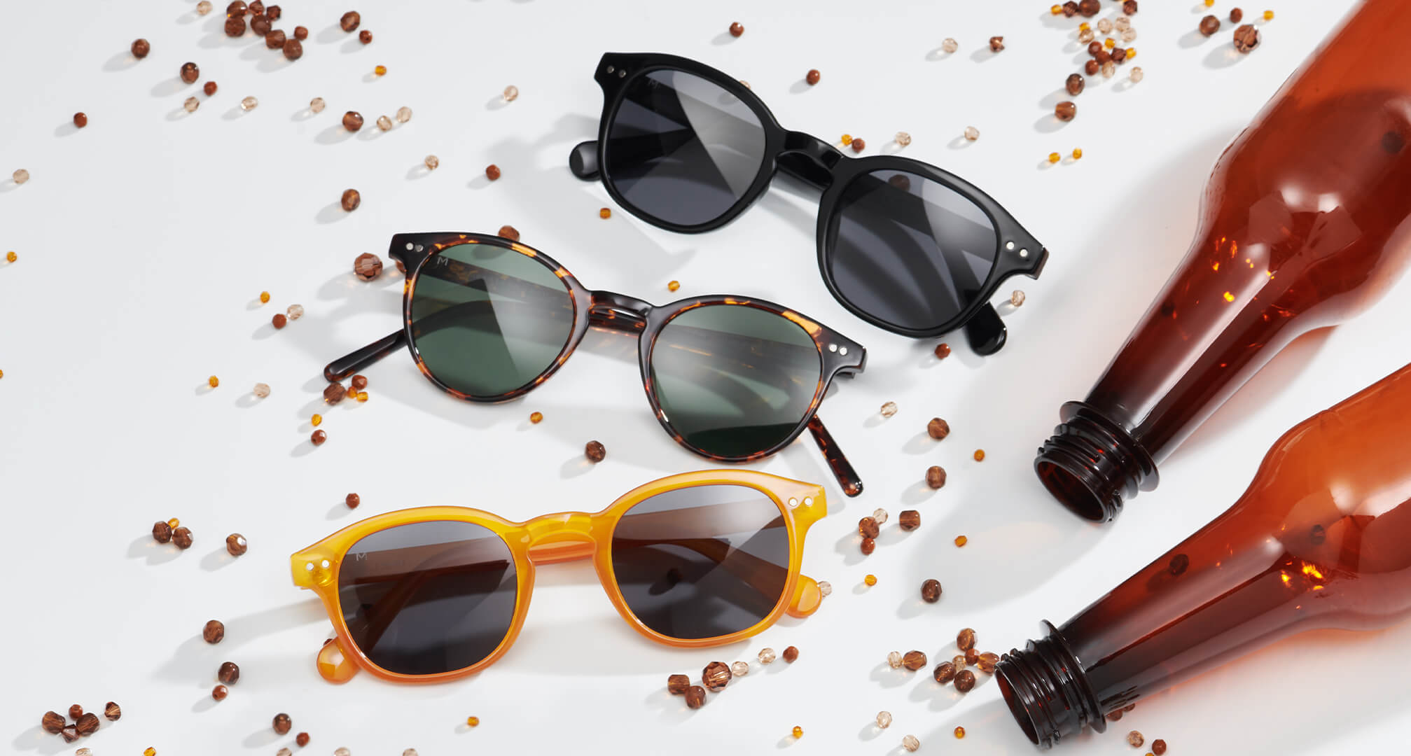 kolme paria aurinkolaseja, jotka on valmistettu kestävistä materiaaleista valkoisella pöydällä, ympärillä helmiä ja muovipulloja.